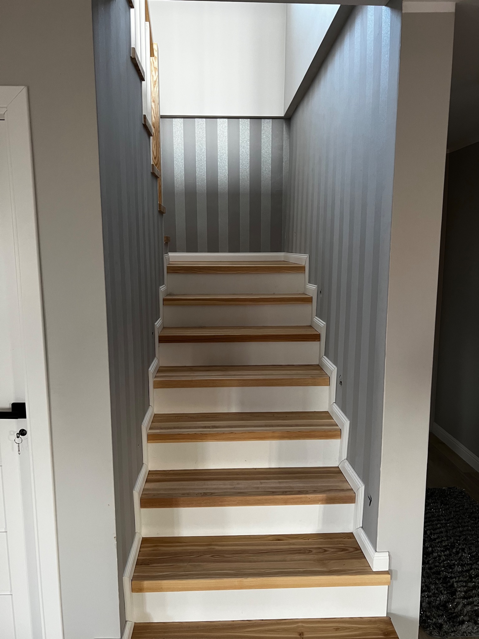 Schody z drewnianymi stopniami i białymi podstopnicami, ściany w odcieniach szarości, pionowe pasy na ścianie w tle.