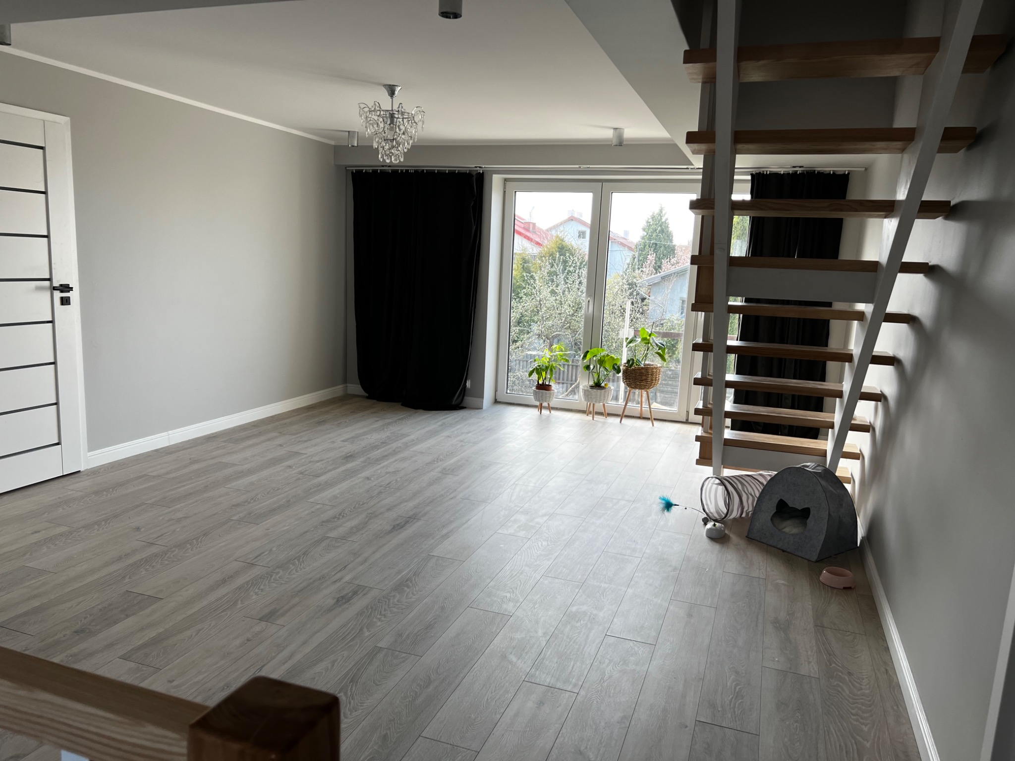 Jasne, przestronne wnętrze po remoncie z szarymi panelami podłogowymi, białymi drzwiami, schodami z drewnianymi stopniami i szarą ścianą, oświetlone przez okno balkonowe z czarnymi zasłonami...