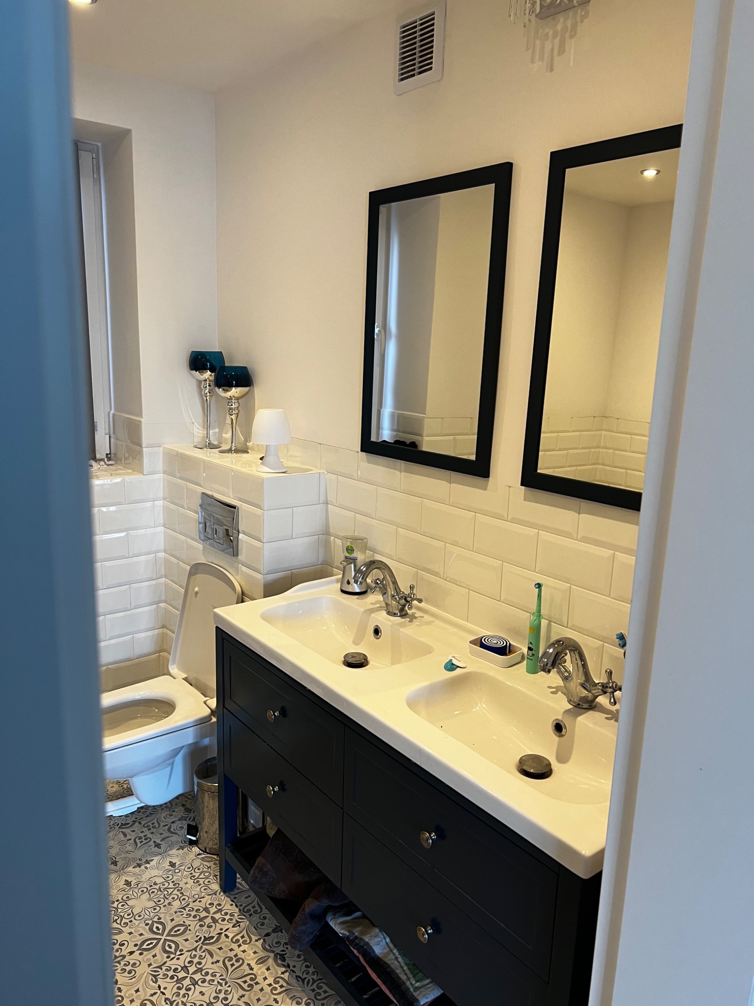 Wykończona łazienka z umywalką z dwiema misami, czarną szafką podumywalkową, lustrami w czarnych ramach i wzorzystą podłogą, widok na toaletę i białe płytki na ścianach.