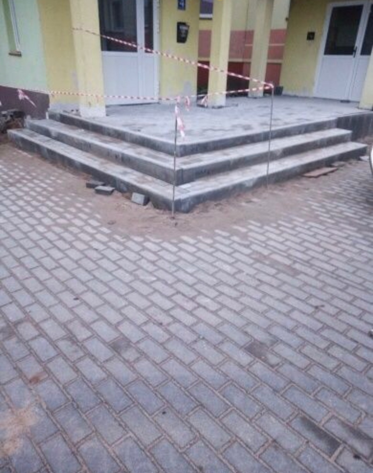 Nowe, szare schody wejściowe z kostki brukowej przed budynkiem, teren zabezpieczony taśmą ostrzegawczą, widoczny fragment ułożonej kostki na chodniku.