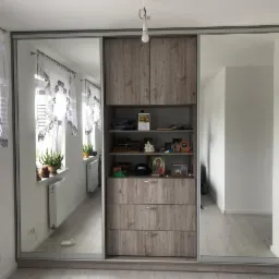 Szafa wnękowa z przesuwnymi lustrzanymi drzwiami, drewnianym korpusem z szufladami i półkami, odbijająca wnętrze jasnego pokoju z roślinami na parapecie.