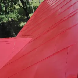 Widok z góry na fragment dachu z blachy w kolorze intensywnej czerwieni, z widocznymi łączeniami paneli i fragmentami zieleni drzew w tle.
