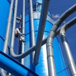 Pracownik w kombinezonie ochronnym i kasku, zabezpieczony linami, maluje przemysłowy silos na tle błękitnego nieba, widoczne liczne rury i instalacje.