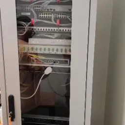 Szafa rack z zamontowanymi urządzeniami sieciowymi: switche, router Cisco, patch panele i zasilacz UPS. Widoczne okablowanie, etykiety na kablach i urządzeniach. Na górze szafy naklejka ostrzegawcza.