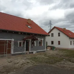 Dwa domy jednorodzinne w r&oacute;żnej fazie prac elewacyjnych: jeden z szarą warstwą izolacyjną i rusztowaniem, drugi z białą elewacją i brązowymi okiennicami, oba z czerwonymi dachami, na tle...