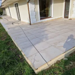 Wyłożony jasnymi płytkami taras przylegający do budynku z kremową elewacją i brązowymi oknami; widoczny przewód od myjki ciśnieniowej na powierzchni tarasu.