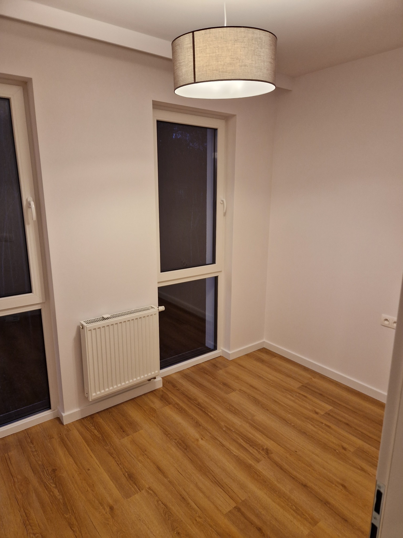 Pusty pokój po remoncie z drewnianą podłogą, białymi ścianami, grzejnikiem pod oknem i wiszącą lampą z abażurem.
