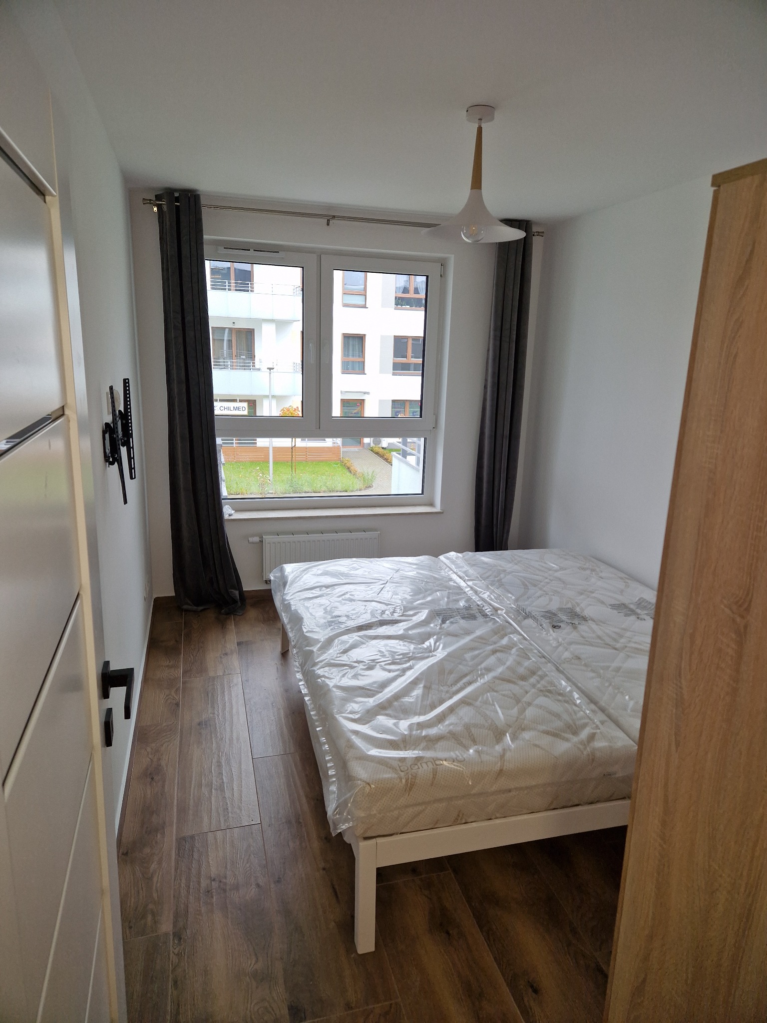 Wnętrze pokoju z drewnianą podłogą, łóżkiem z materacem w folii, grzejnikiem pod oknem i szarymi zasłonami, widok na blok z zielonym trawnikiem.