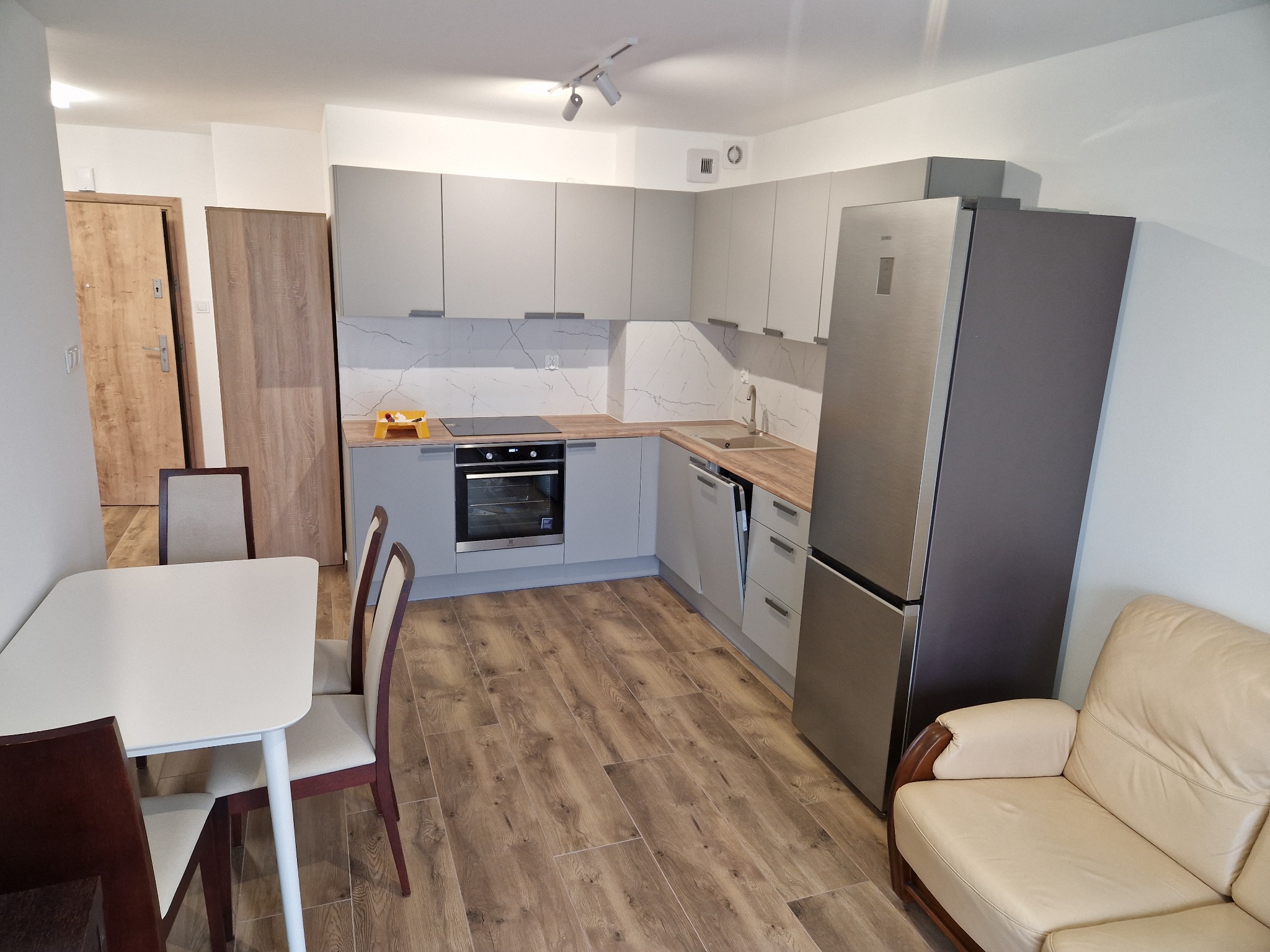 Nowoczesna kuchnia z szarymi szafkami, marmurowym backsplashiem, drewnianym blatem i podłogą, uzupełniona białym stołem z krzesłami i beżową sofą.