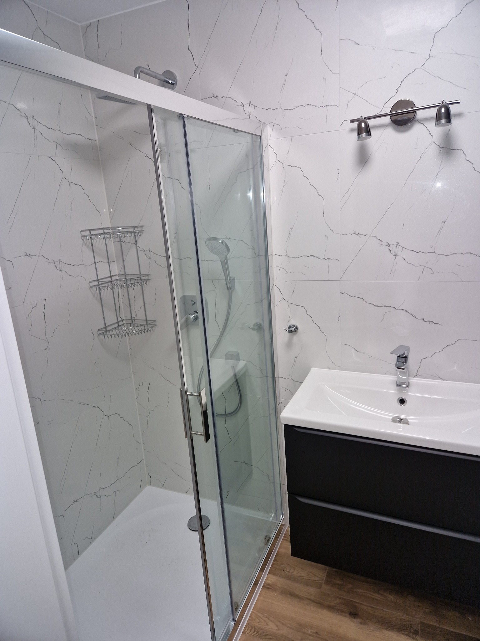 Nowoczesna łazienka z kabiną prysznicową ze szklanymi drzwiami przesuwnymi, umywalką w szafce z czarnymi szufladami i białymi płytkami imitującymi marmur.