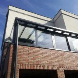 Zadaszenie balkonu z grafitową ramą i szklanym wypełnieniem, osadzone na ceglanej konstrukcji, widok z dołu na tle błękitnego nieba.