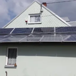 Panele słoneczne zamontowane na dachu domu jednorodzinnego z jasną elewacją i numerem 913, widoczne okno z firanką i ceramiczna ozdoba na parapecie.