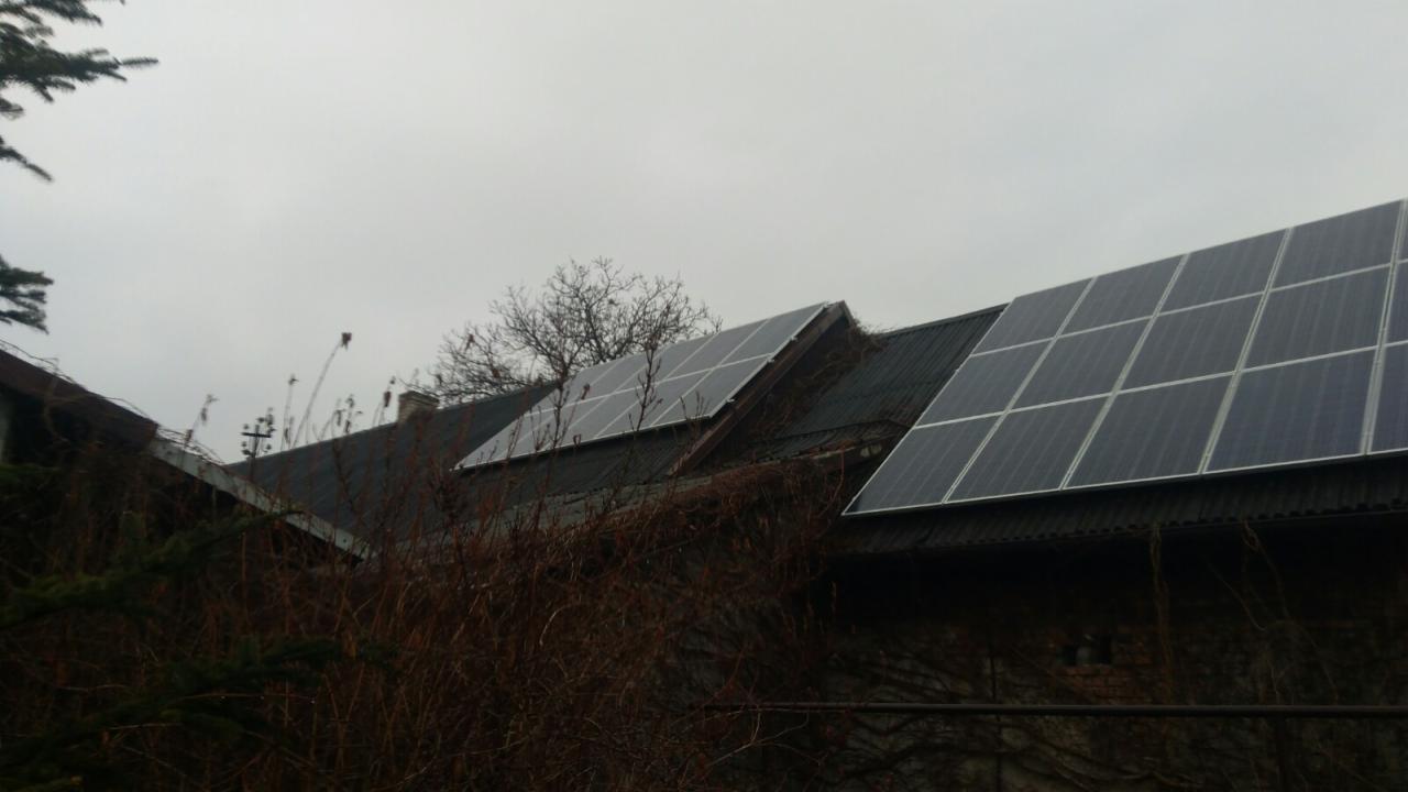 Panele słoneczne zamontowane na dachu starego budynku, widoczne z dołu, z elementami roślinności na pierwszym planie i pochmurnym niebem w tle.