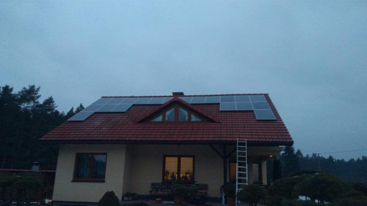 Dom jednorodzinny z czerwoną dachówką, na której zamontowano panele słoneczne; drabina oparta o ścianę budynku. Pochmurne niebo w tle.