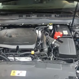 Otwarta maska czarnego samochodu, widoczny silnik Forda z plastikową osłoną, czyste i uporządkowane wnętrze komory silnika, akumulator z czerwonymi zaciskami.