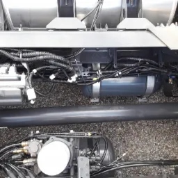 Widok podwozia samochodu z licznymi przewodami, zbiornikiem i elementami mechanicznymi, sfotografowane od dołu na tle żwirowego podłoża.