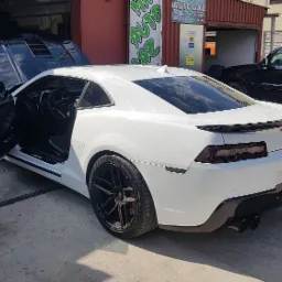 Białe sportowe auto z otwartymi drzwiami na tle warsztatu z szyldem AUTO GAZ i innymi pojazdami.