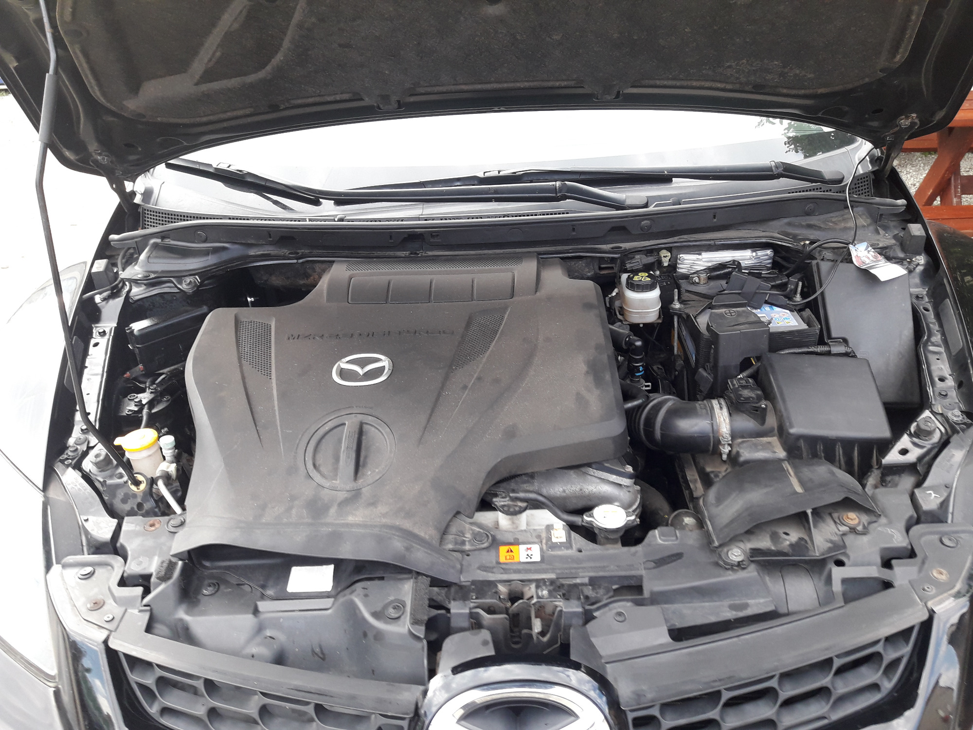 Otwarta maska czarnego samochodu marki Mazda, widoczny zakurzony silnik z elementami osprzętu, w tle fragment drewnianej konstrukcji.