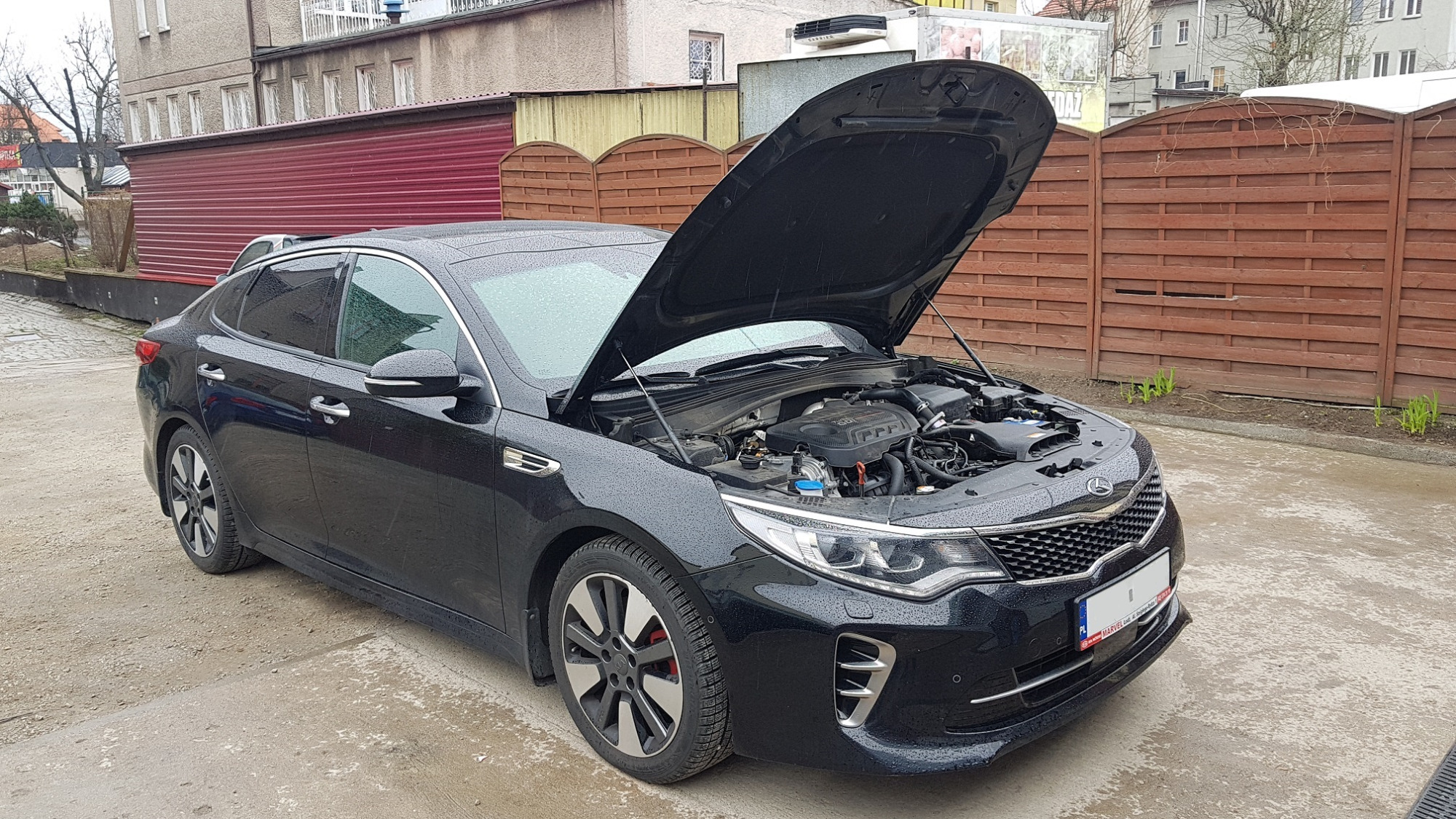 Czarny samochód osobowy z otwartą maską silnika na zewnątrz, widoczny silnik i elementy mechaniczne, tło stanowi budynek i drewniany płot.