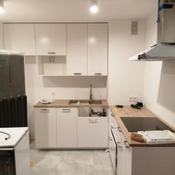 remoncik - Montaż białej, nowoczesnej kuchni z drewnianym blatem, widoczne podłączenia hydrauliczne pod zlewem, płyta indukcyjna w trakcie montażu, narzędzia na blacie.