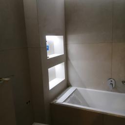 remoncik - Łazienka w trakcie wykańczania, z zabudowaną wanną, wnękami w ścianie wyłożonej szarą płytką i częściowo zakrytym blatem wanny.