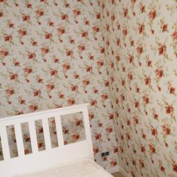 remoncik - Pokój z tapetą w kwiaty, białe łóżko z pościelą, widoczne gniazdka elektryczne na ścianie.