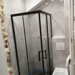 remoncik - Nowoczesna kabina prysznicowa z czarnymi profilami, zamontowana w łazience wykończonej szarymi płytkami. Widoczny fragment ściany z odsłoniętą cegłą i pianką montażową oraz wisząca miska WC...