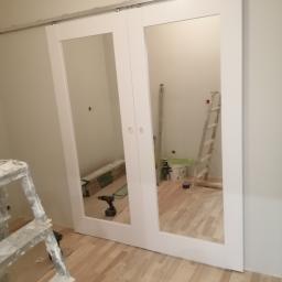 remoncik - Białe, przesuwne drzwi z lustrami podczas montażu, w tle widoczne narzędzia, drabina i panele podłogowe