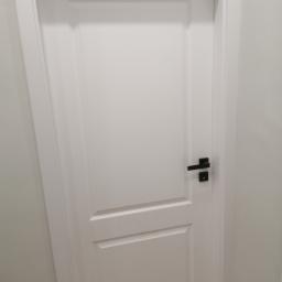 remoncik - Białe, płycinowe drzwi wewnętrzne z czarną, minimalistyczną klamką na tle jasnej ściany.