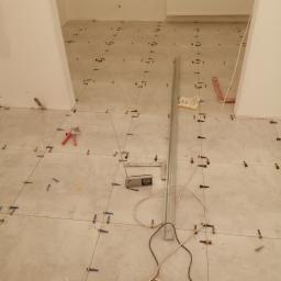 remoncik - Podłoga w trakcie układania płytek z użyciem systemu poziomowania; widoczne krzyżyki dystansowe, poziomica, przedłużacz i butelka z wodą.