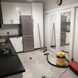 remoncik - Kuchnia w trakcie prac wykończeniowych: nowe, białe szafki kuchenne z czarnym blatem, lodówka w folii ochronnej, na podłodze narzędzia, poziomica, odkurzacz przemysłowy i tuba z klejem.