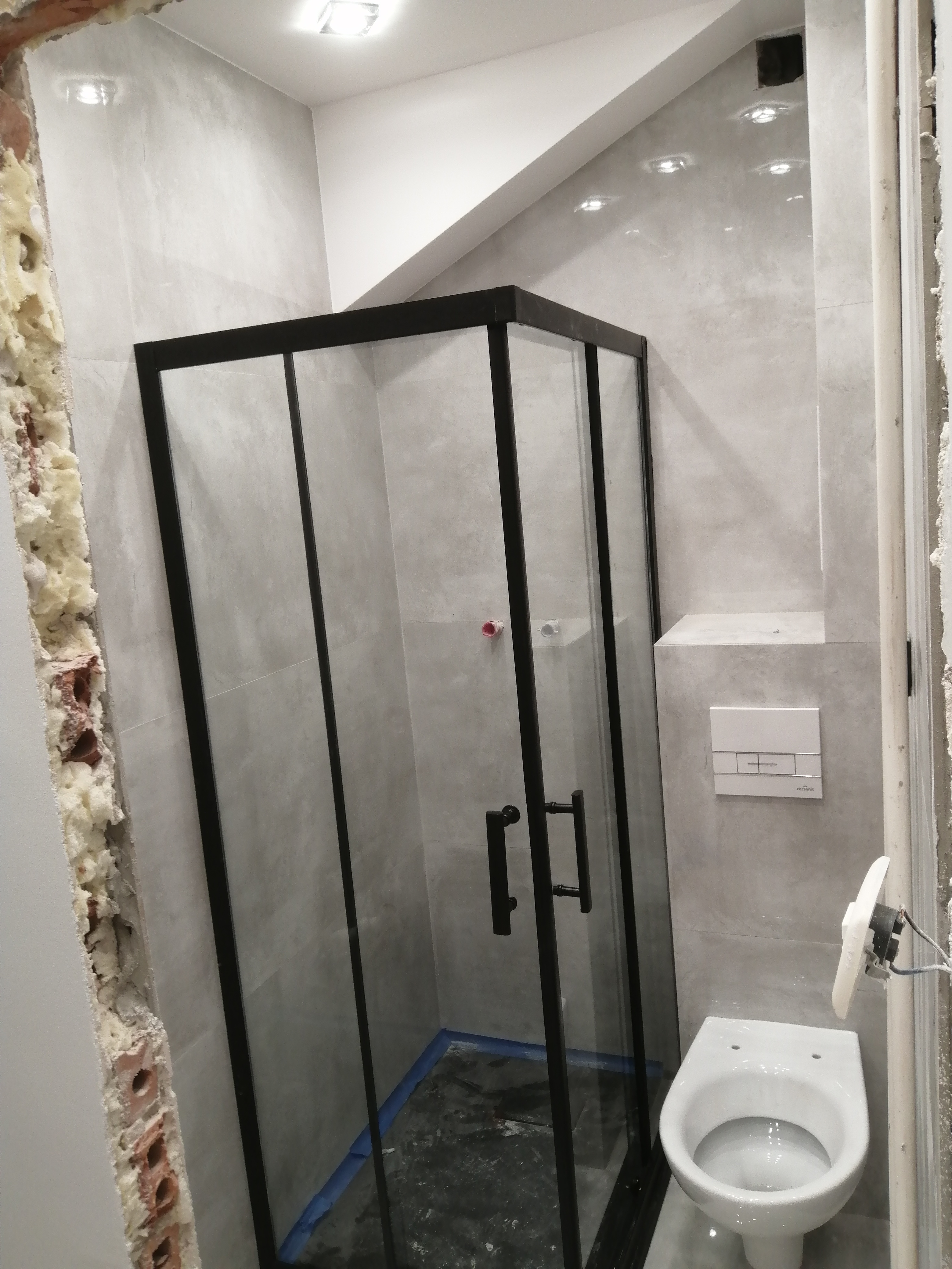 Nowoczesna kabina prysznicowa z czarnymi profilami, zamontowana w łazience wykończonej szarymi płytkami. Widoczny fragment ściany z odsłoniętą cegłą i pianką montażową oraz wisząca miska WC...