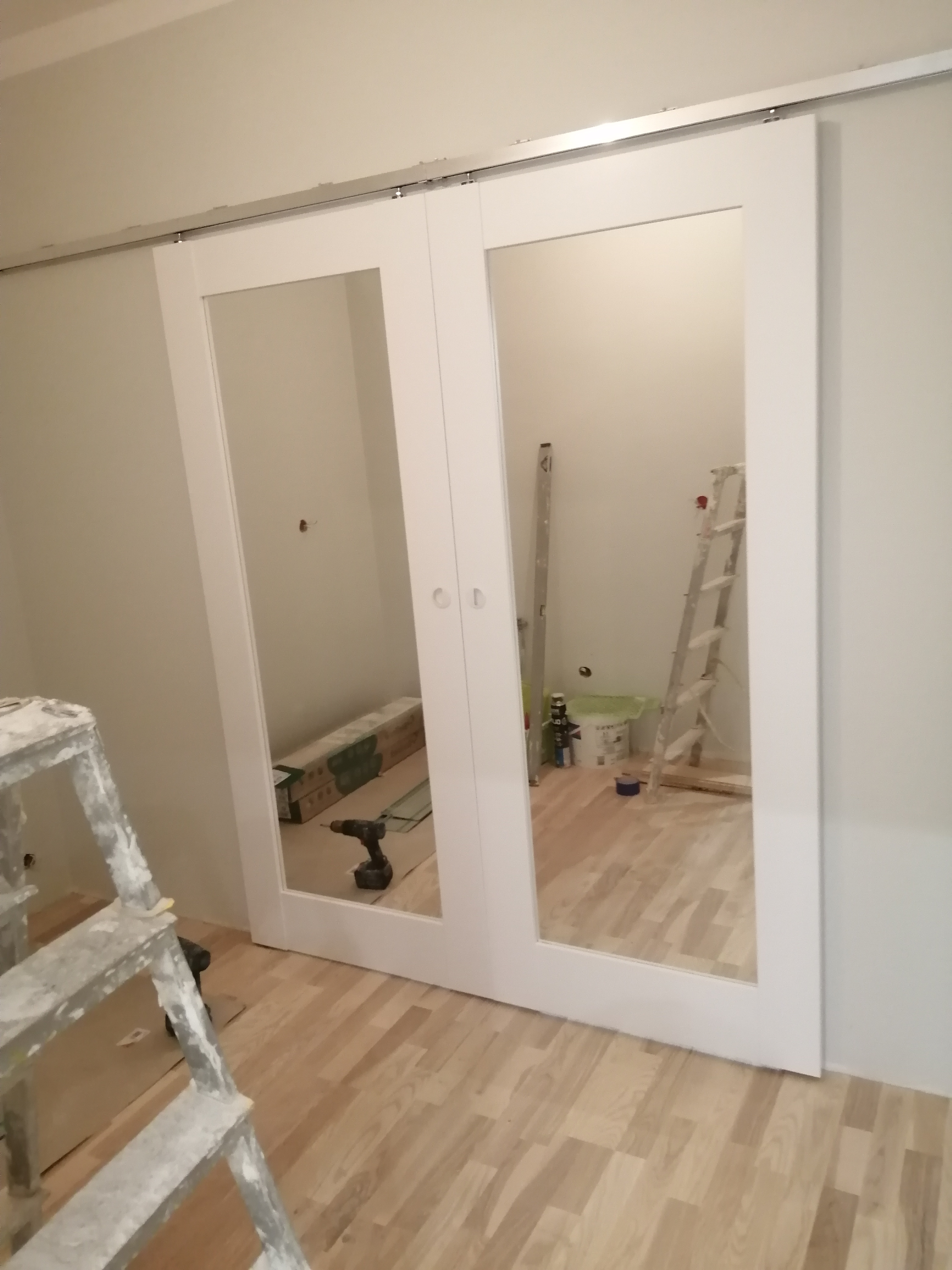 Białe, przesuwne drzwi z lustrami podczas montażu, w tle widoczne narzędzia, drabina i panele podłogowe