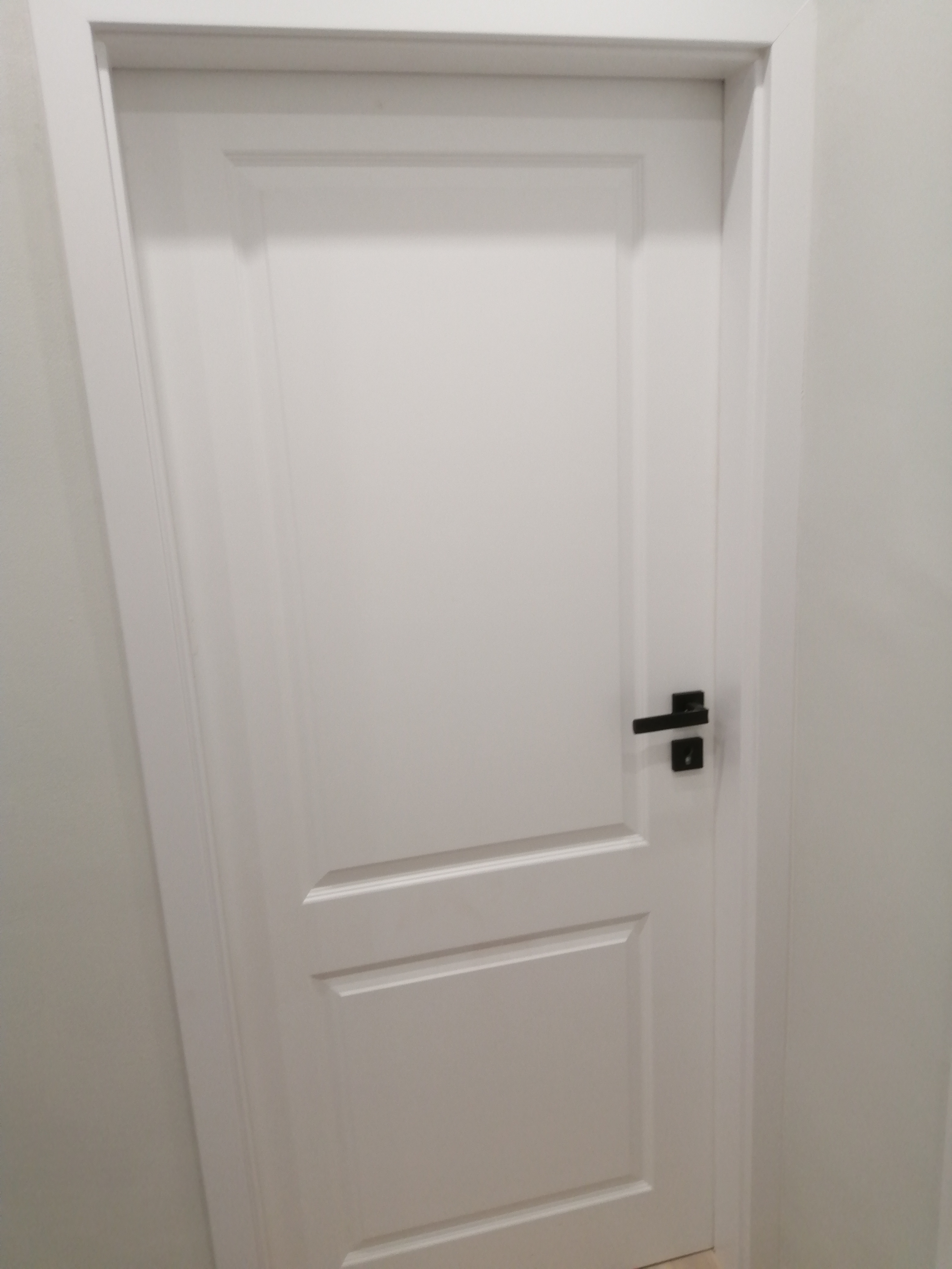 Białe, płycinowe drzwi wewnętrzne z czarną, minimalistyczną klamką na tle jasnej ściany.