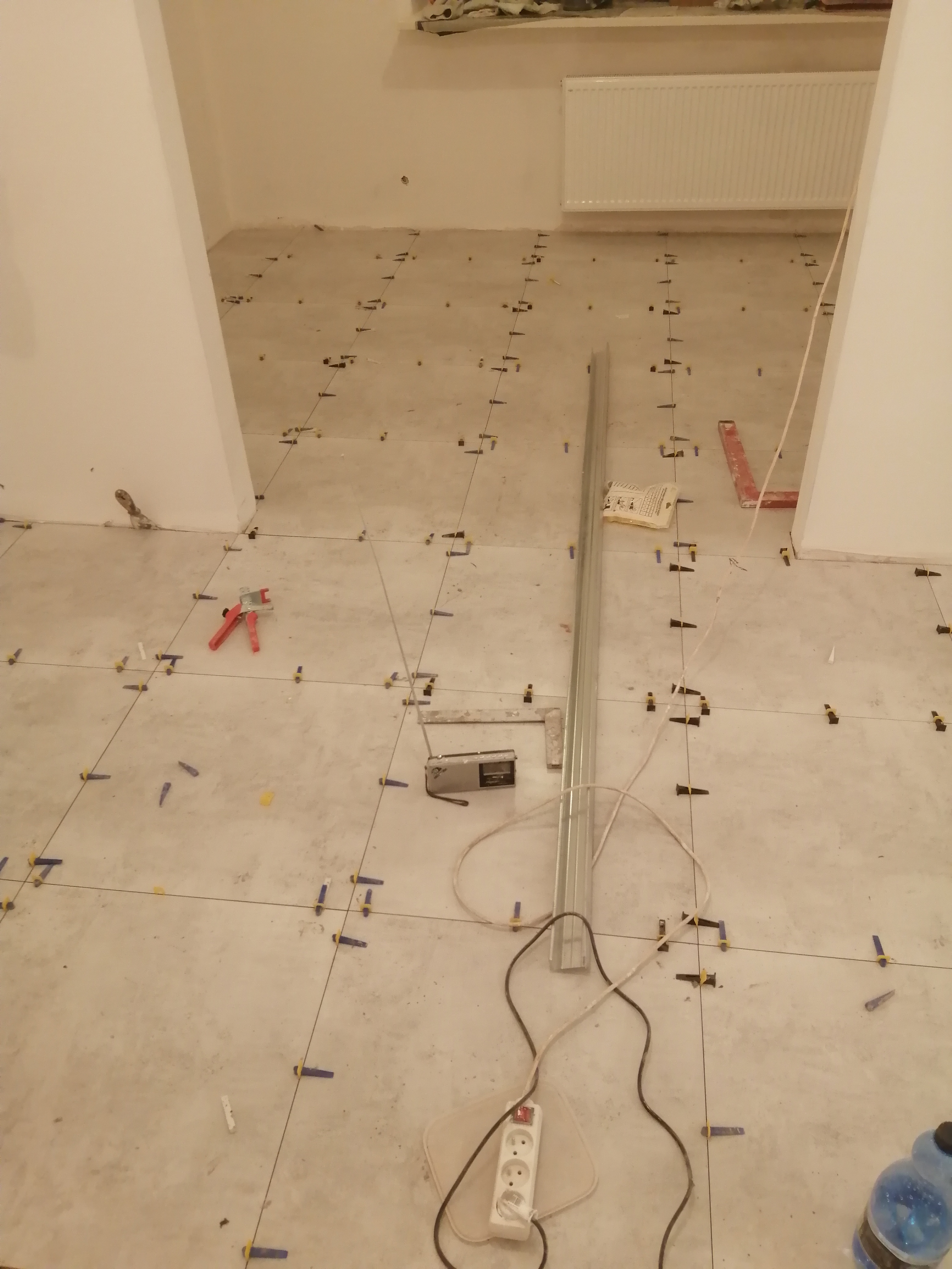 Podłoga w trakcie układania płytek z użyciem systemu poziomowania; widoczne krzyżyki dystansowe, poziomica, przedłużacz i butelka z wodą.