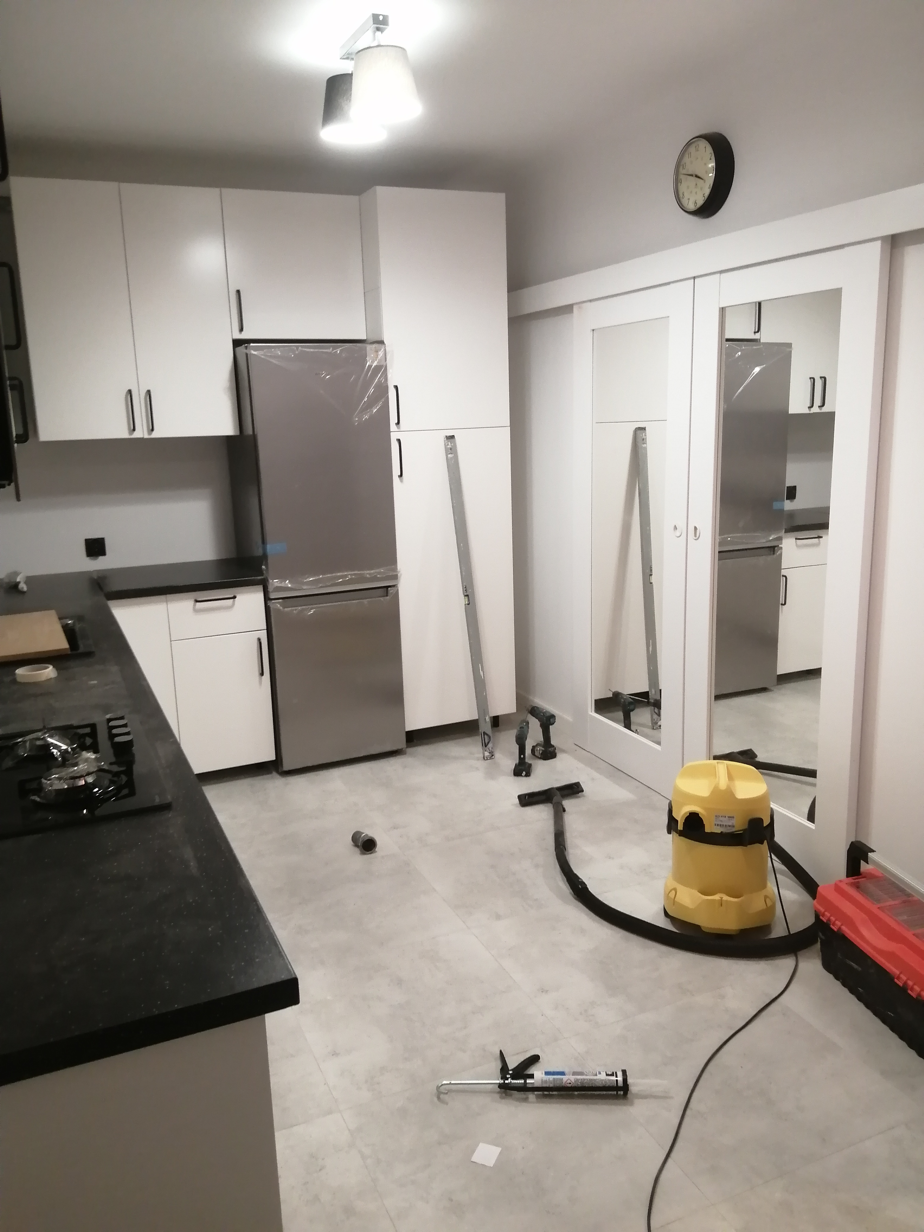 Kuchnia w trakcie prac wykończeniowych: nowe, białe szafki kuchenne z czarnym blatem, lodówka w folii ochronnej, na podłodze narzędzia, poziomica, odkurzacz przemysłowy i tuba z klejem.
