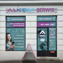 Fasada lokalu z szyldem 'ALKOM SERWIS', oklejona folią reklamową z ofertą skupu, sprzedaży i naprawy telefonów, smartfonów, notebooków i tabletów; widoczna kobieta trzymająca telefon i logotypy...