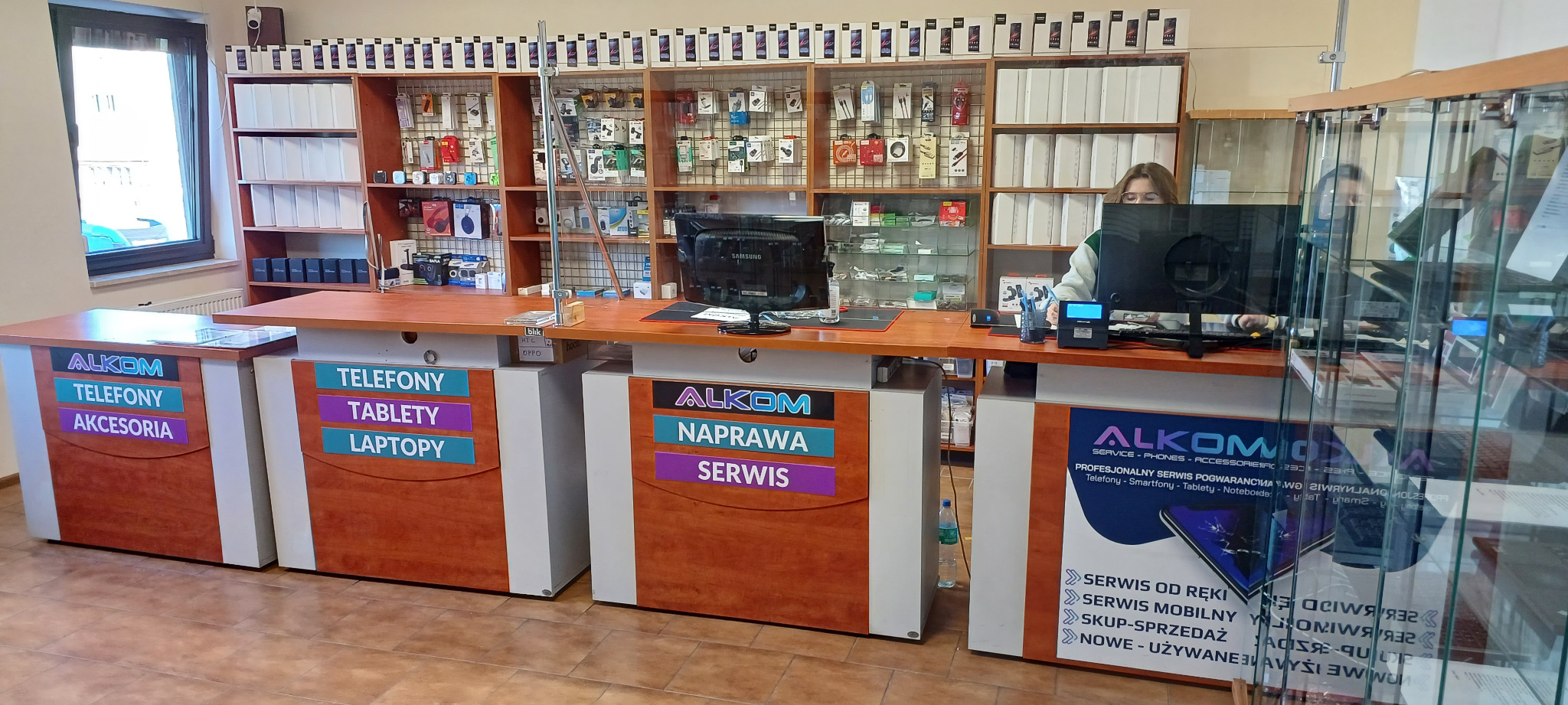 Wnętrze punktu serwisowego telefonów z ladą, za którą stoi pracownik, na półkach akcesoria i telefony, na ladzie monitory i urządzenia serwisowe.