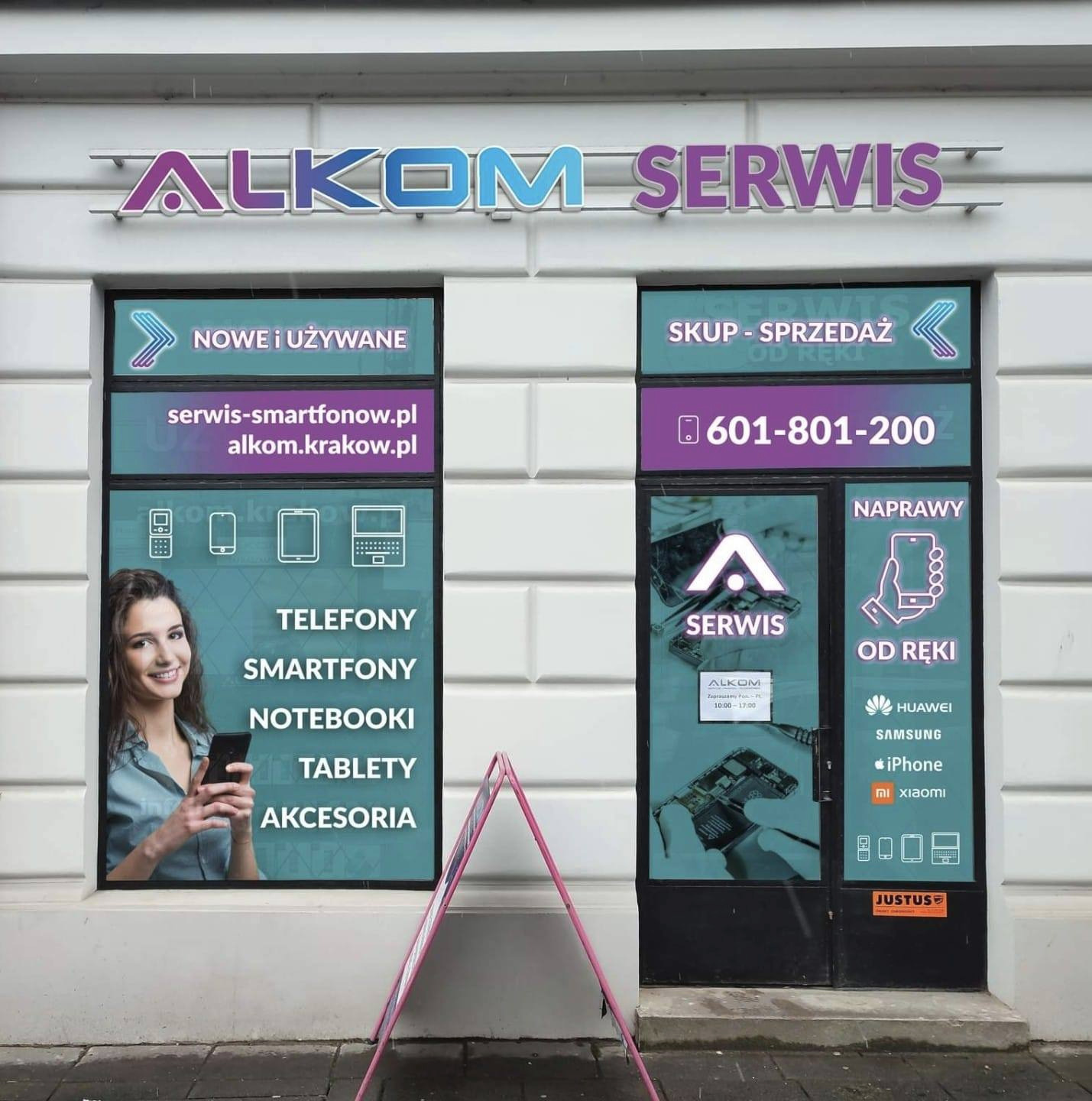 Fasada lokalu z szyldem 'ALKOM SERWIS', oklejona folią reklamową z ofertą skupu, sprzedaży i naprawy telefonów, smartfonów, notebooków i tabletów; widoczna kobieta trzymająca telefon i logotypy...