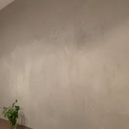 Ściana pokryta dekoracyjnym tynkiem w odcieniach szarości, z widoczną fakturą i subtelnymi nierównościami, obok apteczka pierwszej pomocy i roślina w szklanym wazonie.