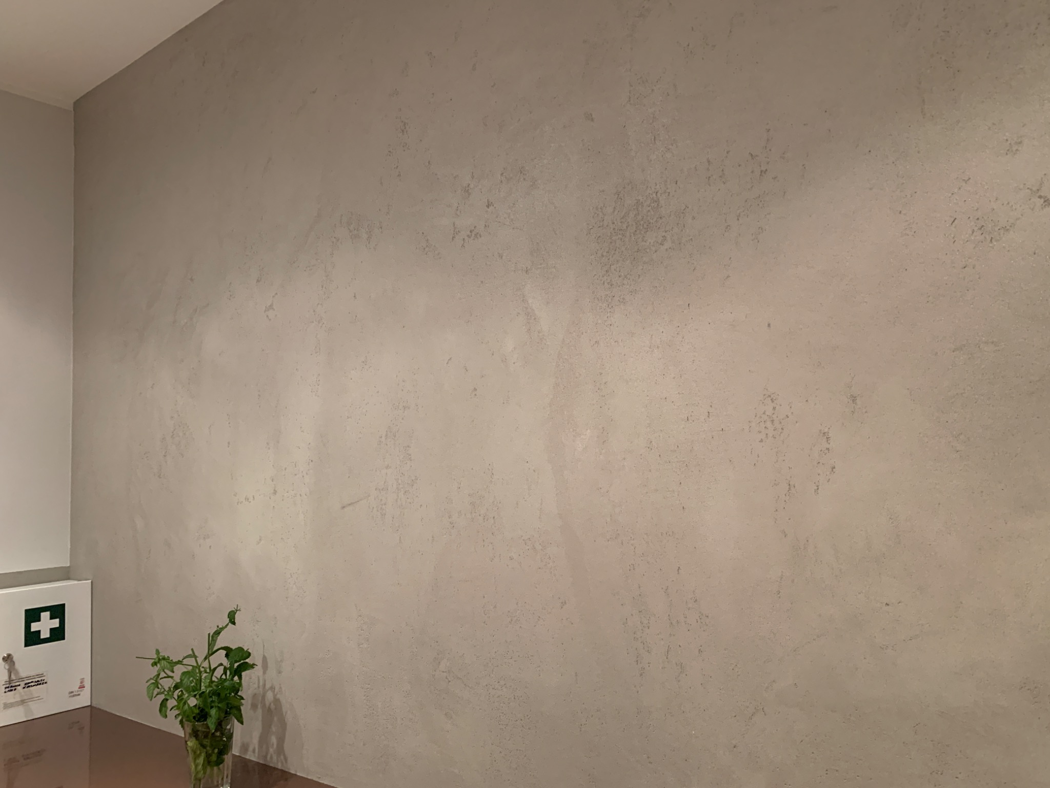 Ściana pokryta dekoracyjnym tynkiem w odcieniach szarości, z widoczną fakturą i subtelnymi nierównościami, obok apteczka pierwszej pomocy i roślina w szklanym wazonie.