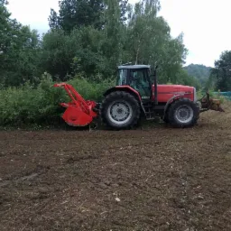 Czerwony traktor z frezem mulczującym, pracujący na polu z resztkami roślinności, w tle drzewa i krzewy.