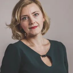 Portret kobiety z blond włosami, ubrana w ciemnozieloną sukienkę z ozdobnym wycięciem, uśmiechnięta z delikatnym makijażem.