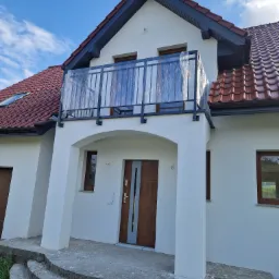 Nowy balkon z czarną, metalową konstrukcją zabezpieczony folią ochronną, widok na biały dom z czerwoną dachówką i błękitne niebo.