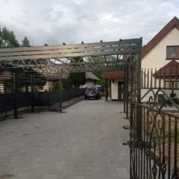 Metalowa konstrukcja zadaszenia nad podjazdem z widoczną bramą kutą i fragmentem domu w tle.