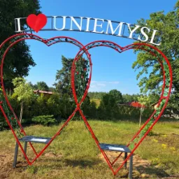 Dwa czerwone, metalowe serca tworzące bramę/pergole z miejscami do siedzenia, ozdobione zielonymi elementami imitującymi pnącza i czerwonymi kwiatami, z napisem 'I ❤️ UNIEMYŚL' u góry, na tle...