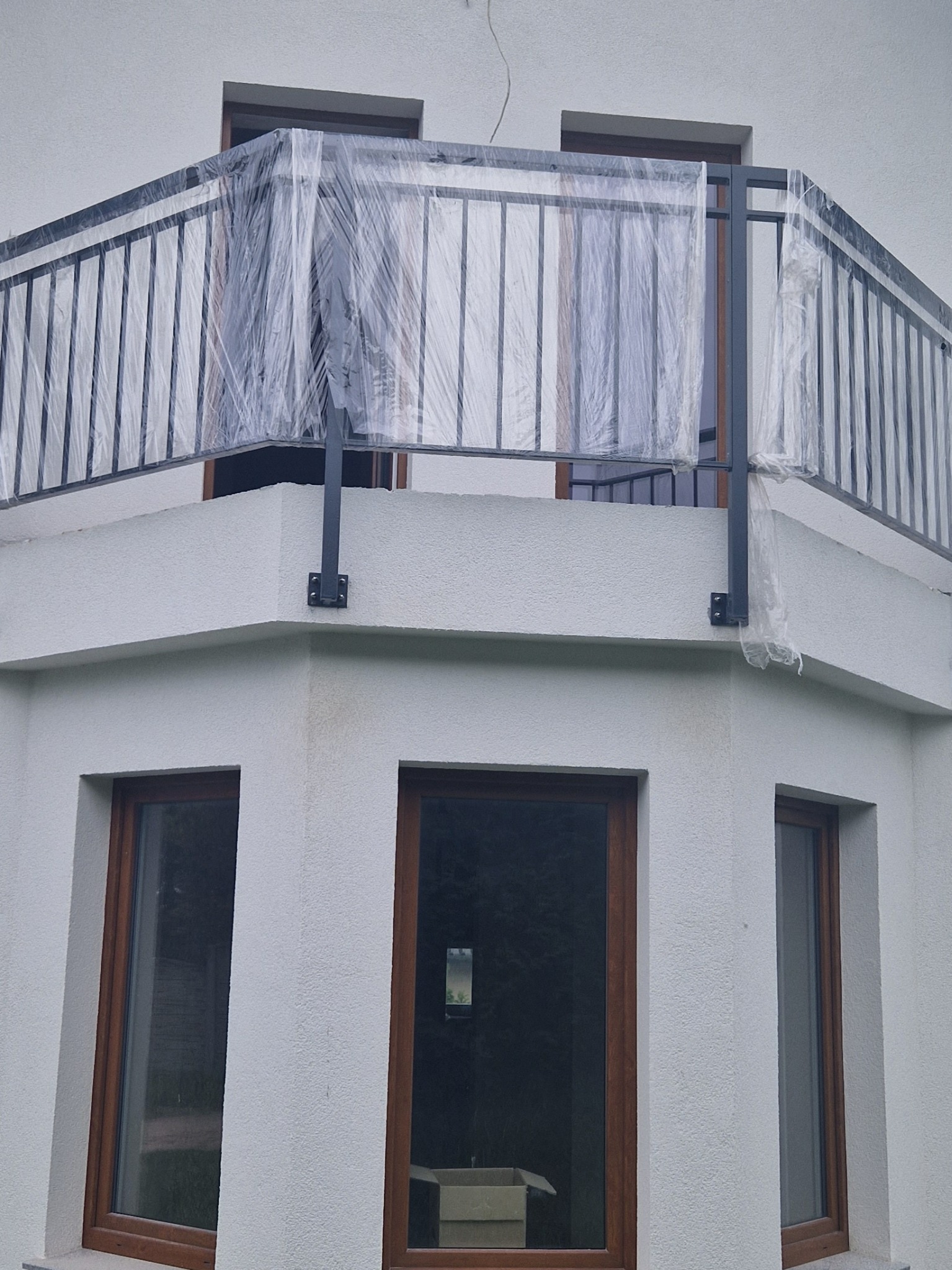 Czarna metalowa balustrada balkonowa, zabezpieczona folią ochronną podczas prac budowlanych, widoczna z zewnątrz budynku z drewnianymi oknami.