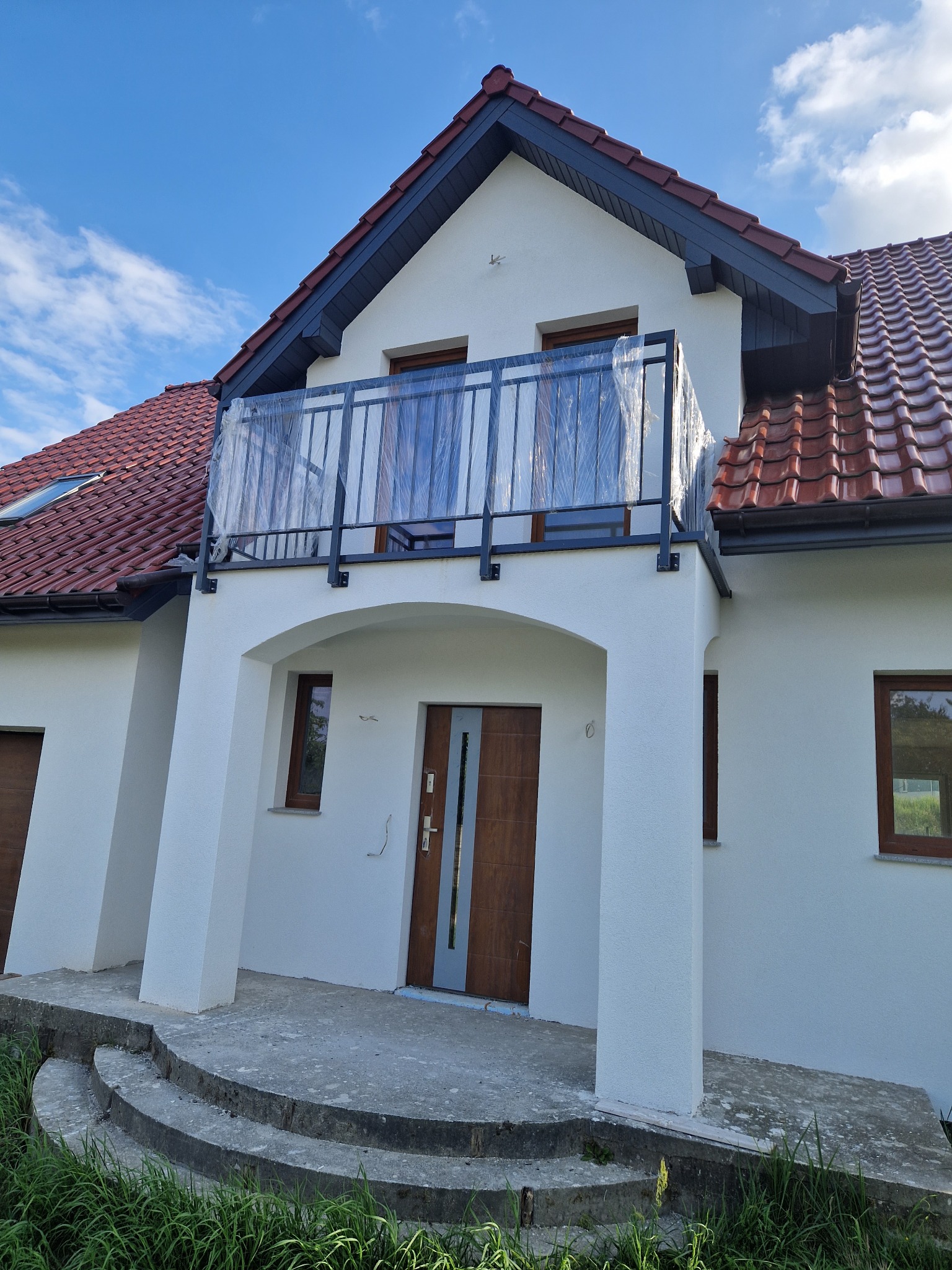 Nowy balkon z czarną, metalową konstrukcją zabezpieczony folią ochronną, widok na biały dom z czerwoną dachówką i błękitne niebo.