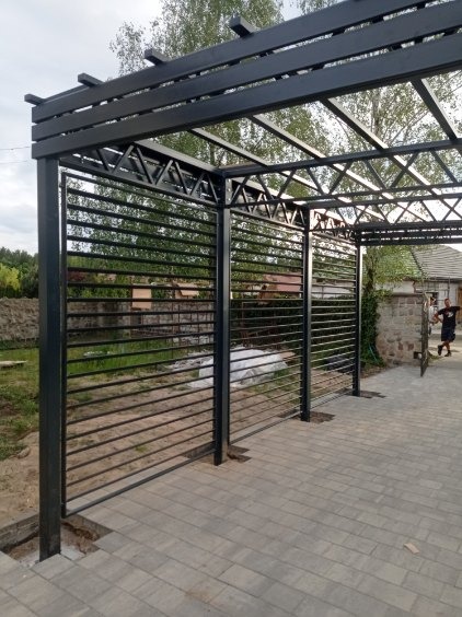 Czarna, metalowa pergola z poziomymi listwami, widok z boku, na kostce brukowej, w tle zieleń i budynek.
