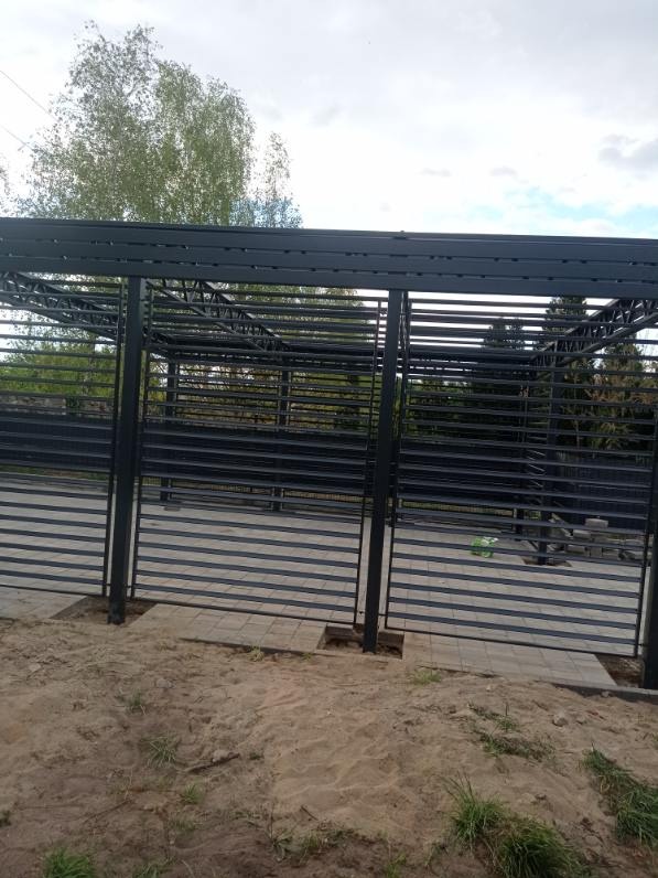 Konstrukcja metalowa, prawdopodobnie zadaszenie lub wiata, w trakcie montażu, widoczne fundamenty i kostka brukowa wokół słupów, tło stanowi drzewo i pochmurne niebo.